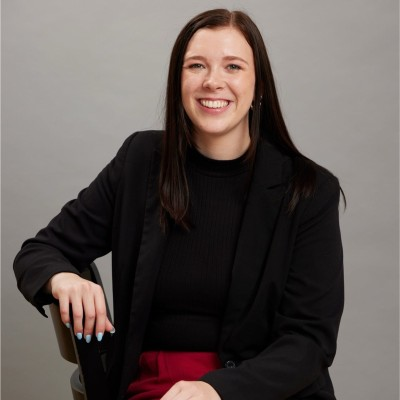 Hannah St. Clair sits in a chair wearing a black blazer and black top with red pants. Her left arm rests on the chair and her right hand is on her lap. She has long dark hair and light blue nail polish. The background is a neutral gray studio setting. The overall tone is professional and approachable. There is no visible text in the image.